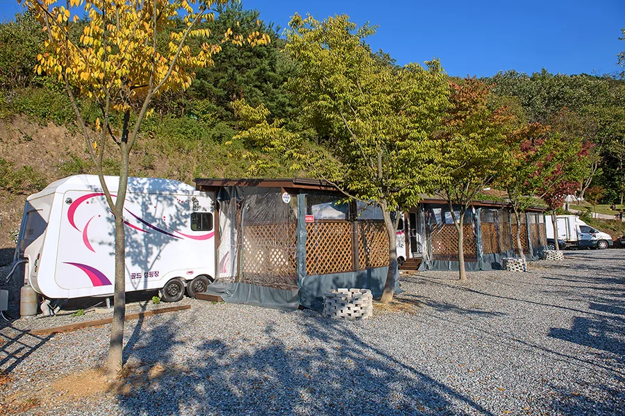 비탈 아래 자갈 마당에 늘어선 카라반과 차양 시설, 뒤편의 숲